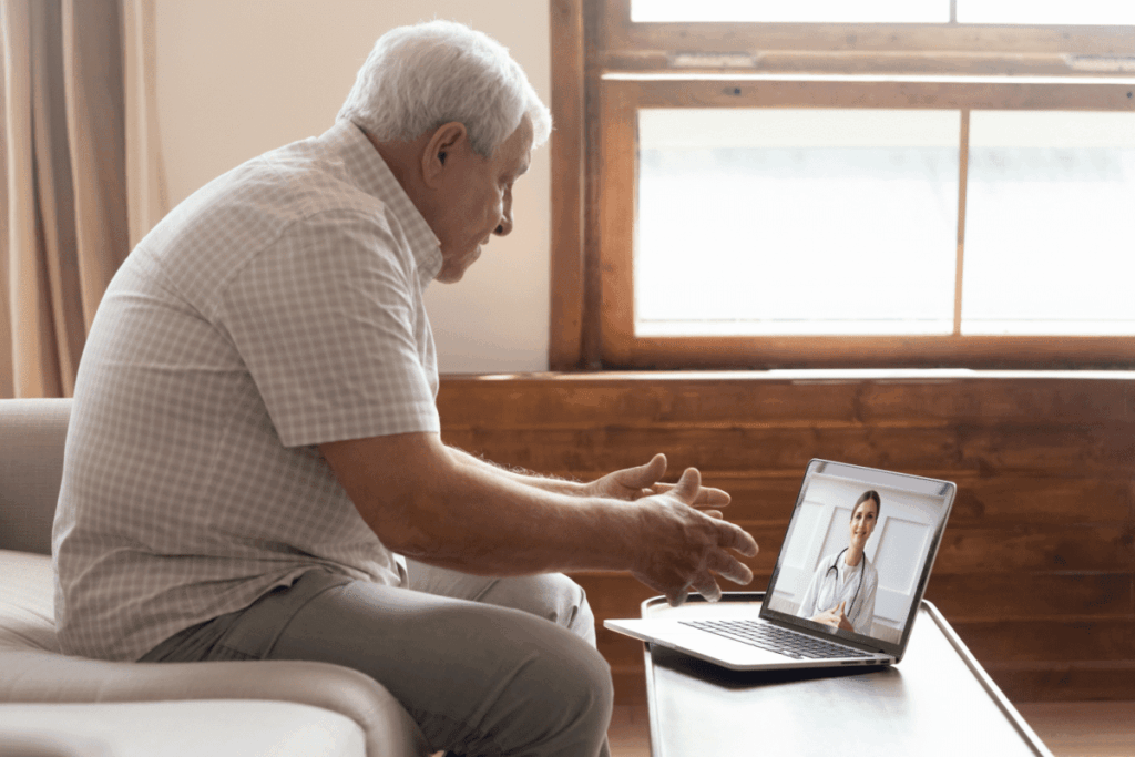Older man seated on a home sofa, gesturing toward a laptop showing a smiling female doctor in a white coat.