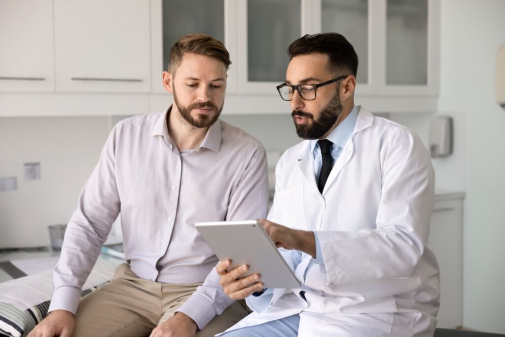 Male doctor in a white coat showing a tablet to a seated male patient in a clinical exam room.