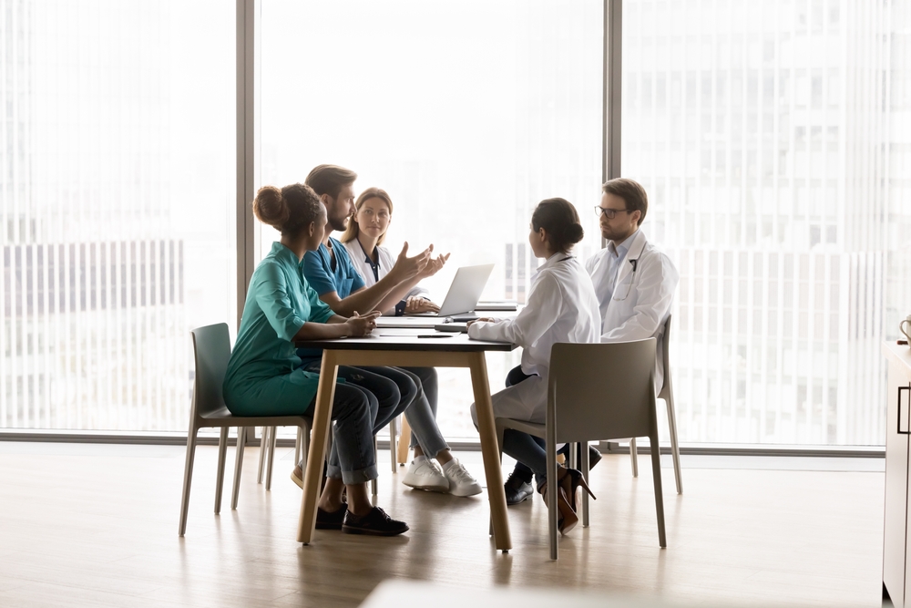 Healthcare professionals in scrubs and white coats meet around a conference table in a bright, windowed office.