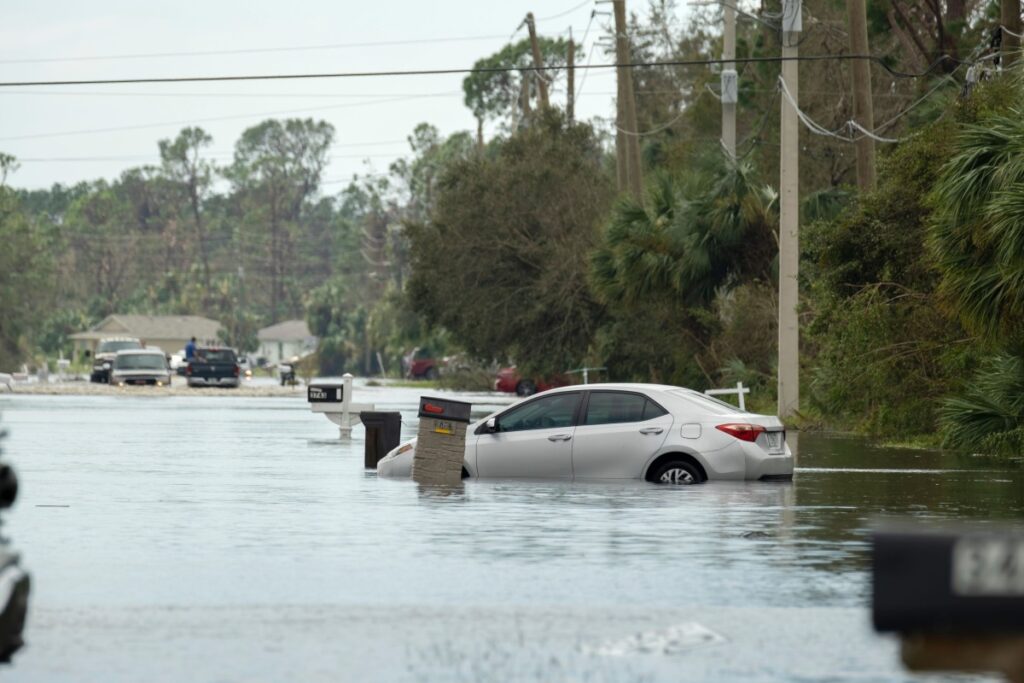 Flooded residential street with partially submerged cars, mailboxes, utility poles and standing water after a storm.